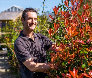 Rob Crowle wearing Duchy of Cornwall Nursery uniform and smiling while standing beside a red plant.